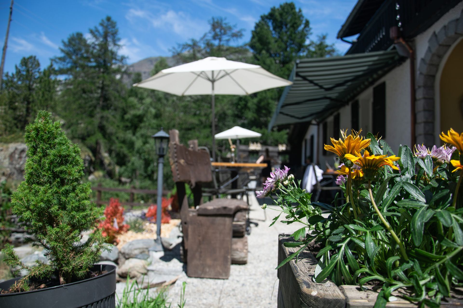 Sonnenterrasse des Restaurant Alm in Furi: Sonnenschirm, Holztische und Blumen vor dem Bergpanorama